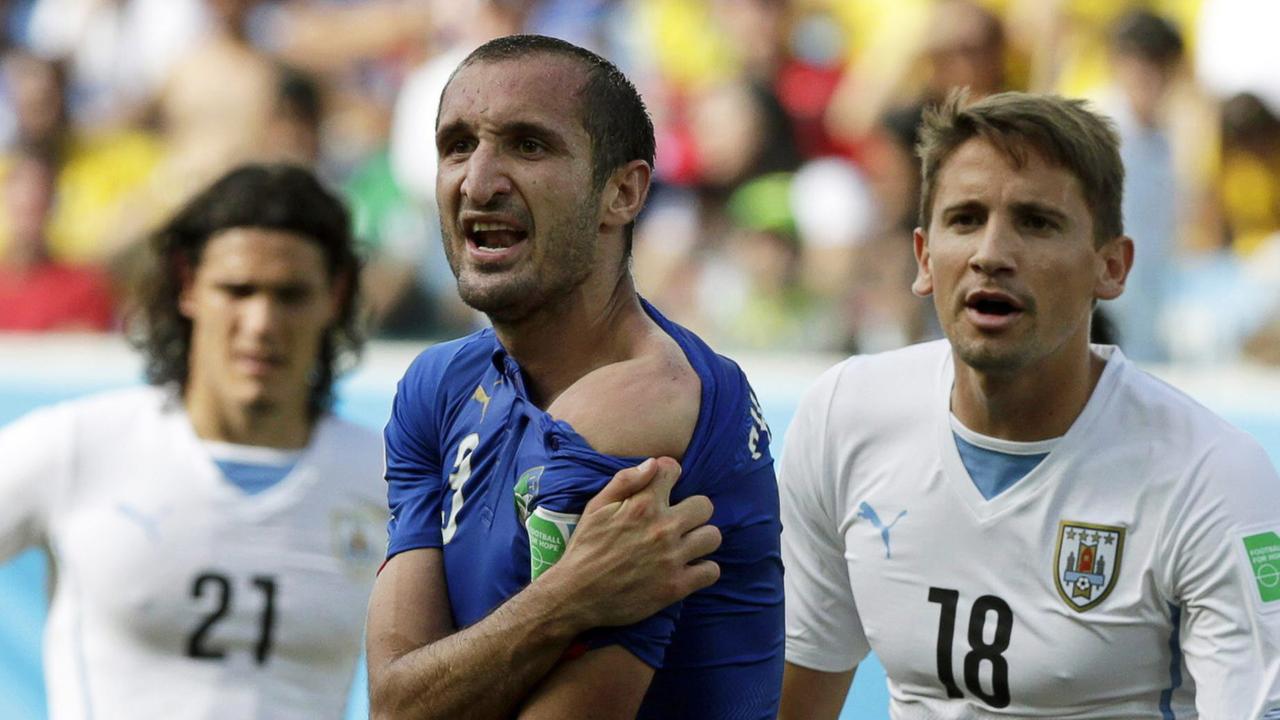 Giorgio Chiellini zieht sein Trikot über seine linke Schulter herunter, um Spuren des angeblichen Bisses zu zeigen, links und rechts hinter ihm zwei Spieler aus Uruguay