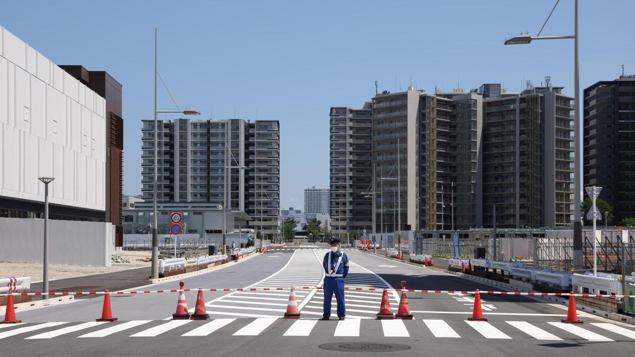 Blick auf die Hochhaussiedlung des olympischen Dorfes in Tokio. Im Bildvordergrund ist eine Staßenabsperrung und ein Wachmann mit Schutzmaske zu sehen. Blick auf die Hochhaussiedlung des olympischen Dorfes in Tokio. Im Bildvordergrund ist eine Staßenabsperrung und ein Wachmann mit Schutzmaske zu sehen.