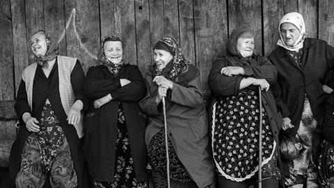 Alte bulgarische Frauen stehen in traditioneller Kleidung und lachend vor einer Holzwand.
