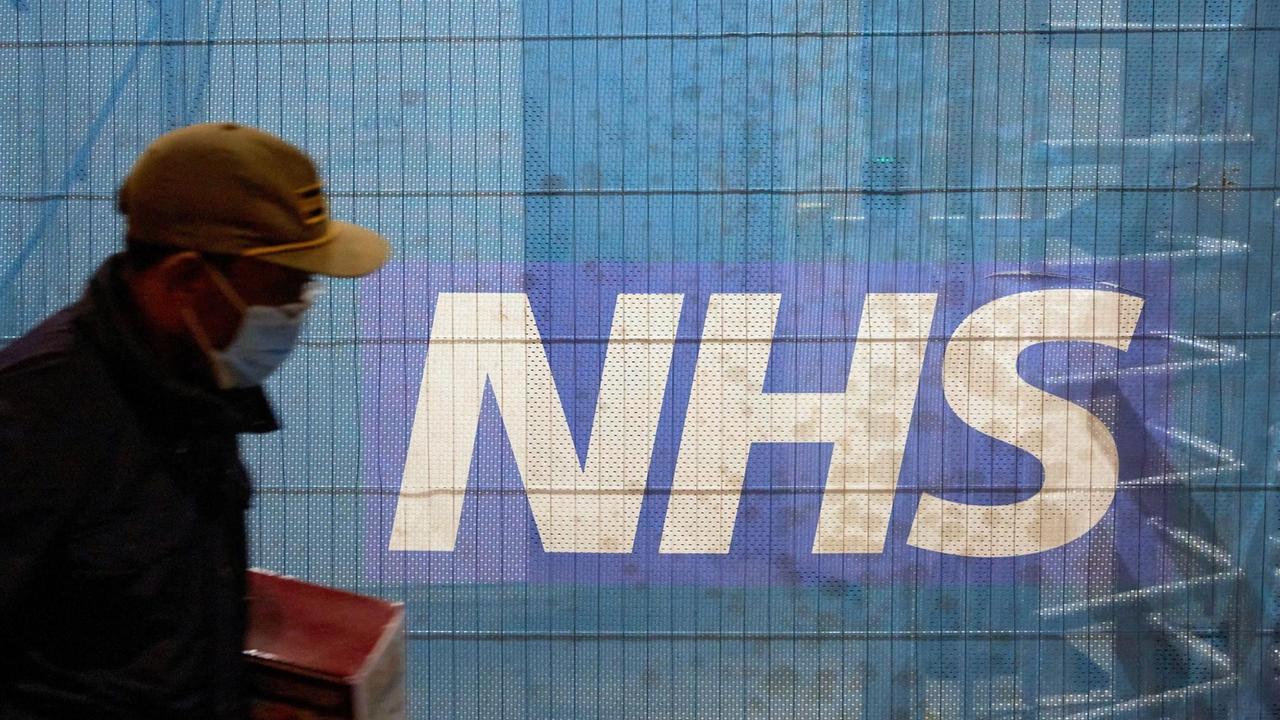 Ein Fußgänger mit einer maske vor einem Banner mit der Aufschrift NHS. Ein Fußgänger mit einer maske vor einem Banner mit der Aufschrift NHS.