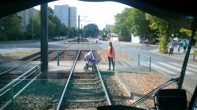 Eine Frau mit oranger Warnweste hat ihre Hand an einem leeren Kinderwagen, der auf Straßenbahnschienen steht.