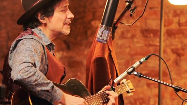 Ein Mann mit Hut auf dem Kopf sitzt Gitarre spielend vor einem Mikrofon. Ein Mann mit Hut auf dem Kopf sitzt Gitarre spielend vor einem Mikrofon.