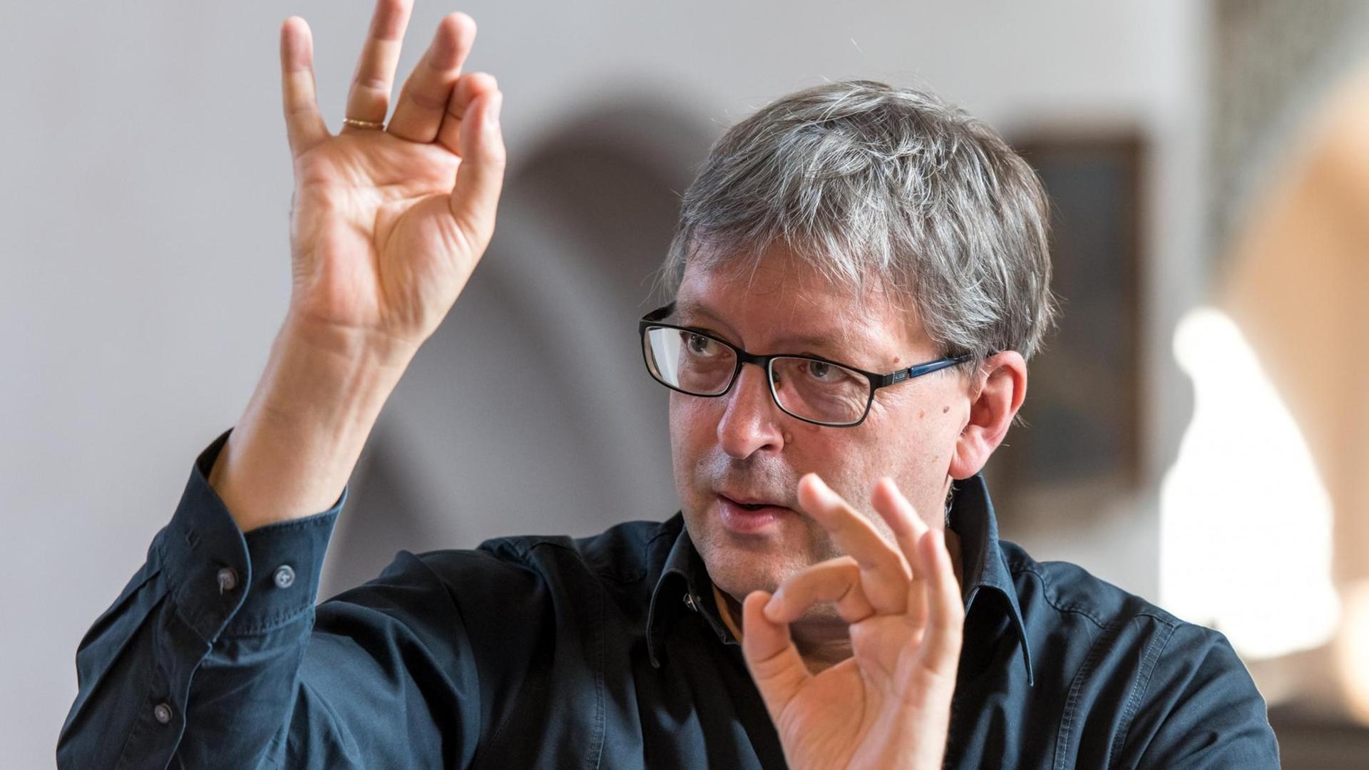 Ein Mann mit Brille und kurzen grauen Haaren hält die Hände in Dirigierposition vor sein konzentriertes Gesicht. Ein Mann mit Brille und kurzen grauen Haaren hält die Hände in Dirigierposition vor sein konzentriertes Gesicht.