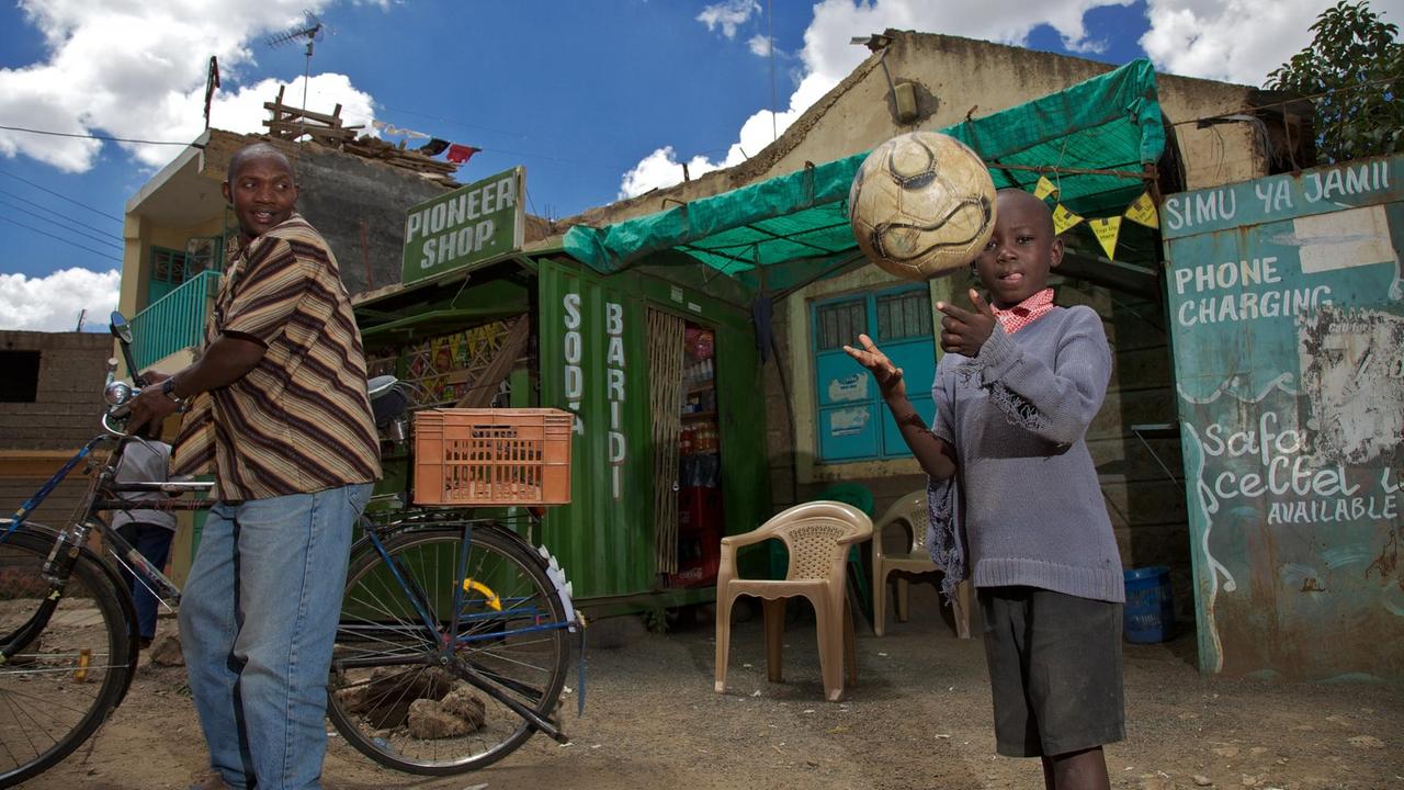 In einer Straße in Mathare wirft ein kleiner Junge einen Fußball in die Luft. Dahinter sind ärmliche Hütten zu sehen.