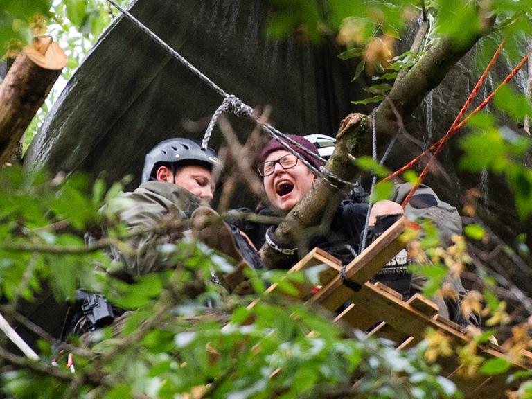 Das Bild ist von unten aufgenommen: Man sieht durch die Blätter den Kopf einer schreienden Umweltaktivistin und eines Polizisten mit Helm.