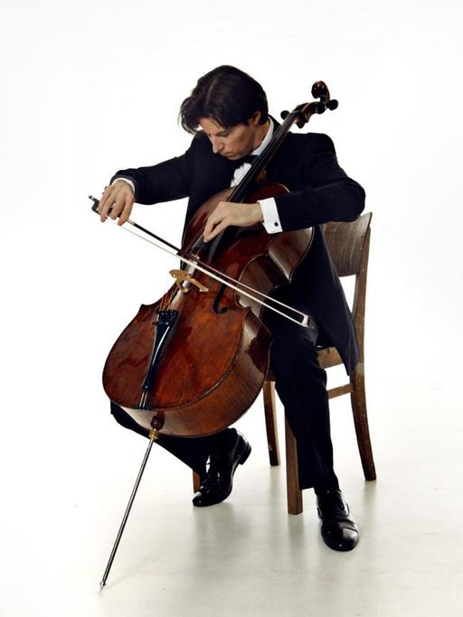 Ein Mann in Anzug sitzt mit seinem Cello in einem weißen Raum