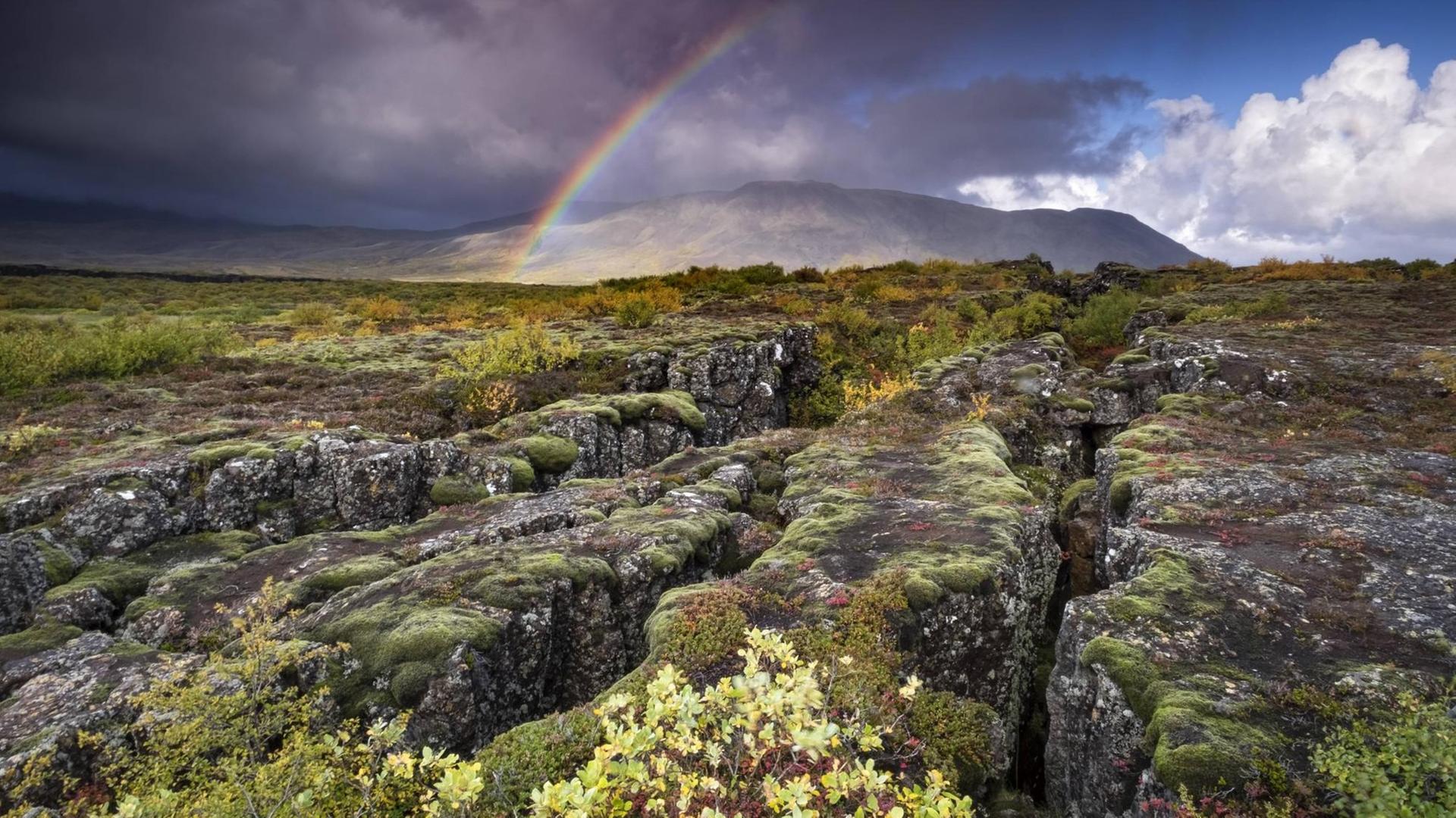 Regenbogen und Regenwolken über einer Vulkanlandschaft mit tektonischer Plattenfissur.