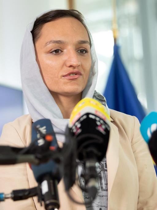 Eine Frau mit selbstbewusstem Blick und Kopftuch spricht in Fernsehmikrofone.