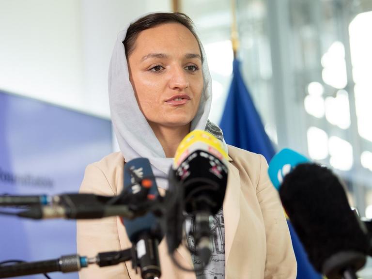 Eine Frau mit selbstbewusstem Blick und Kopftuch spricht in Fernsehmikrofone.