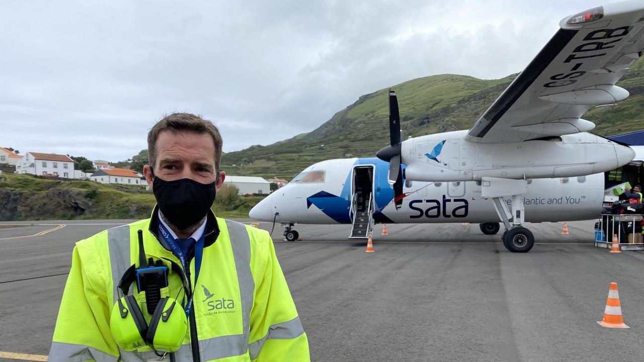 Ein Mann mit gelber Signaljacke und Mundschutz steht auf dem Flughafenfeld. Im Hintergrund ein Flugzeug. Ein Mann mit gelber Signaljacke und Mundschutz steht auf dem Flughafenfeld. Im Hintergrund ein Flugzeug.