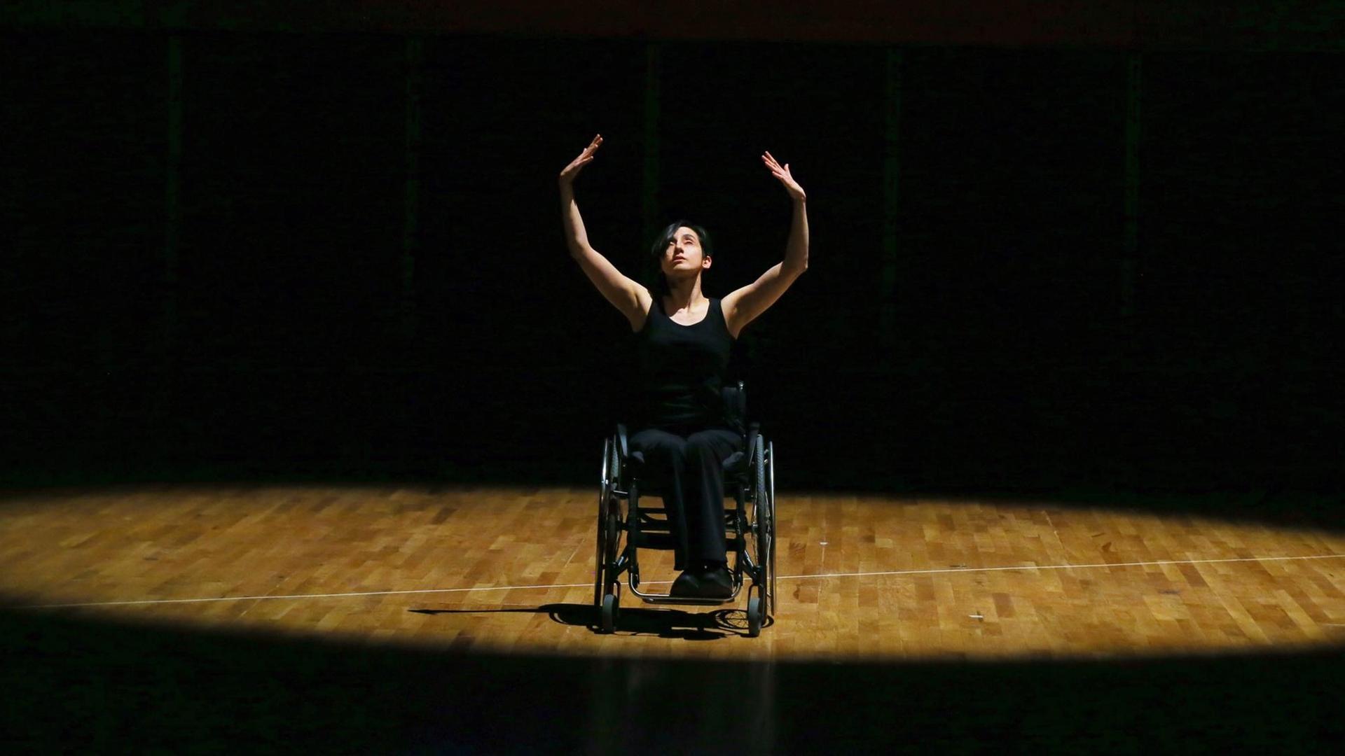 Eine Tänzerin im Rollstuhl performt im Lichtkegel einer sonst dunklen Bühne und streckt ihre Hände in die Höhe. Eine Tänzerin im Rollstuhl performt im Lichtkegel einer sonst dunklen Bühne und streckt ihre Hände in die Höhe.