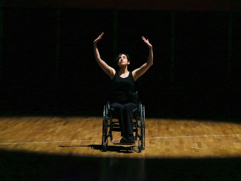 Eine Tänzerin im Rollstuhl performt im Lichtkegel einer sonst dunklen Bühne und streckt ihre Hände in die Höhe.