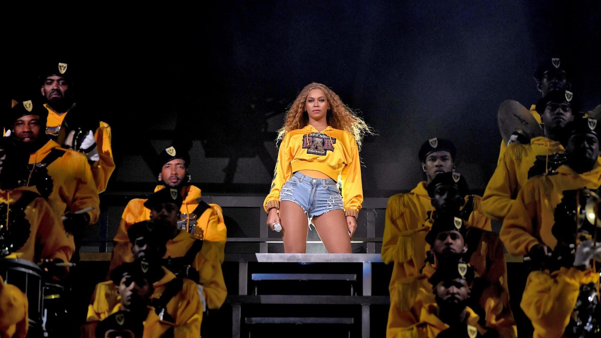 Beyoncé steht in abgerissenen Jeans und einem knallgelben Pullover auf der Bühne und schaut ins Publikum. Beyoncé steht in abgerissenen Jeans und einem knallgelben Pullover auf der Bühne und schaut ins Publikum.