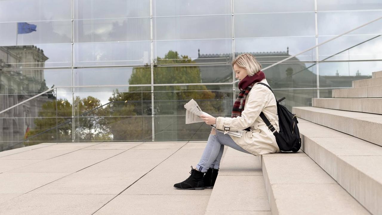 Eine junge Frau sitzt auf einer Betontreppe vor einer Glaswand und liest in einer Zeitung