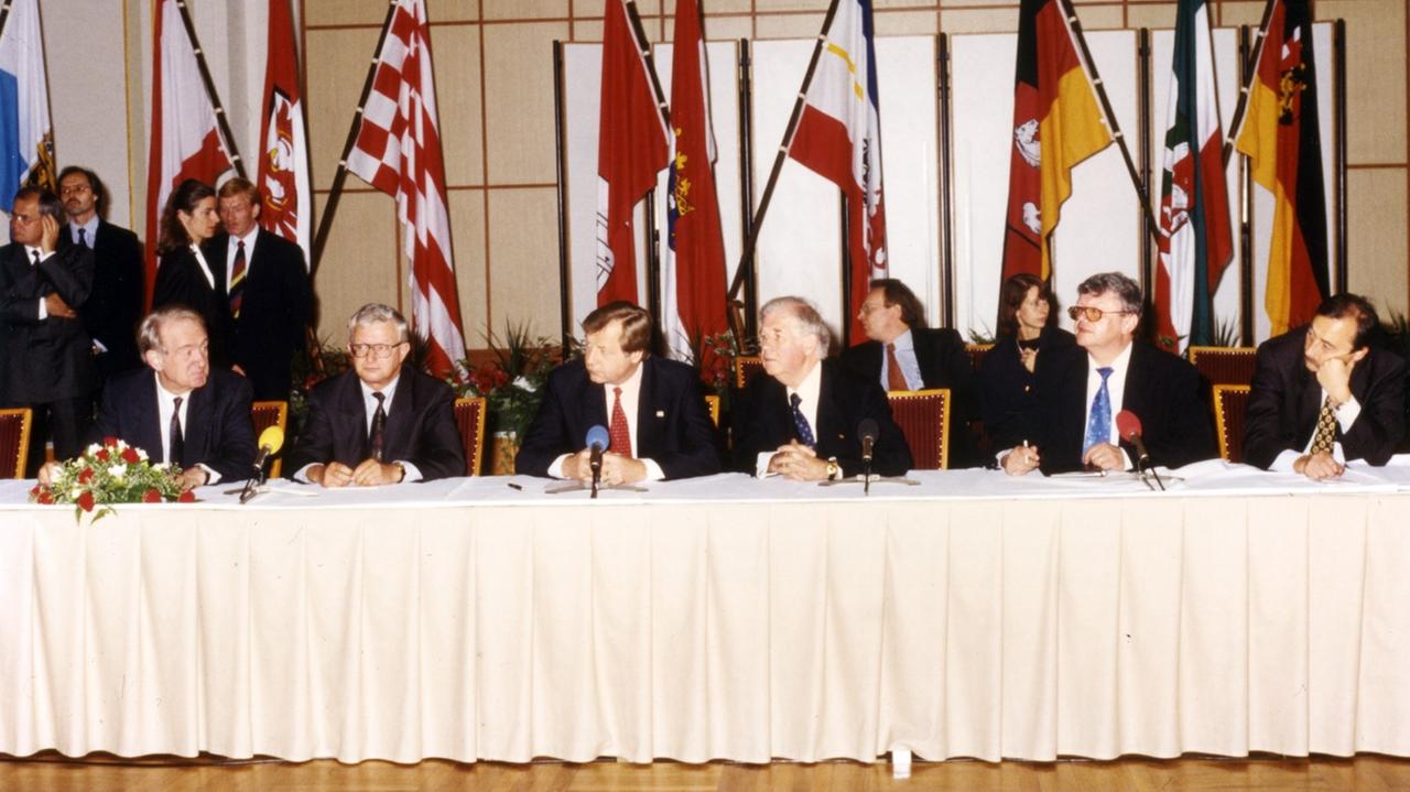 Die Unterzeichnung der Staatsverträge für die Gründung von Deutschlandradio. Im Hintergrund sind Fahnen verschiedener Bundesländer zu sehen.