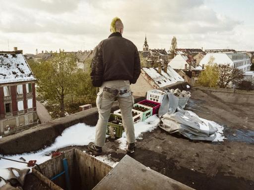 Ein junger Mann mit Irokesenschnitt steht auf dem Dach eines besetzten Hauses und schaut auf das Leipziger Szeneviertel Connewitz.