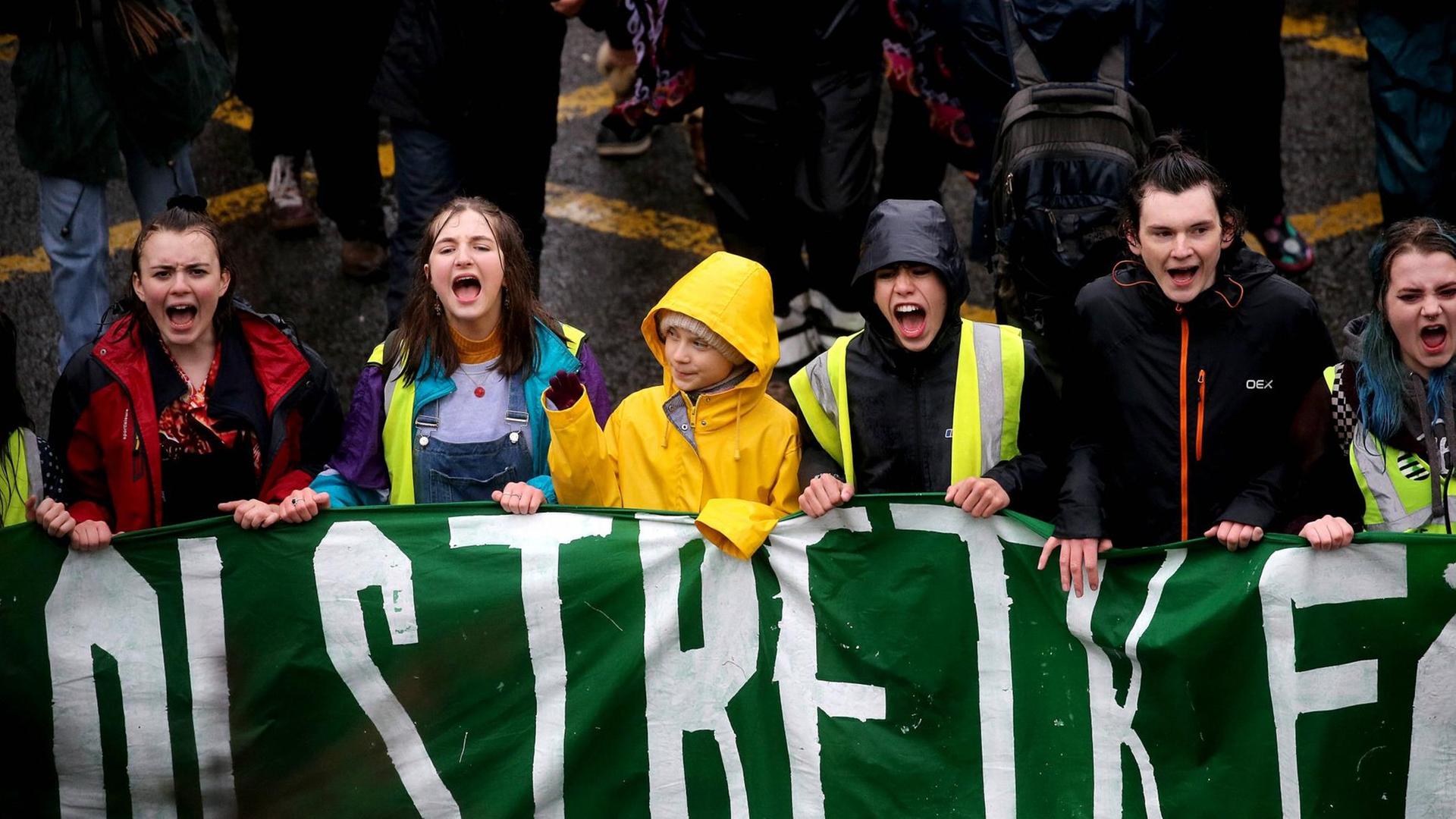 Greta Thunberg hält mit jungen Demonstrierenden ein grünes Banner. Die anderen Jugendlichen rufen etwa. Thunberg schaut zur Seite und winkt. Greta Thunberg hält mit jungen Demonstrierenden ein grünes Banner. Die anderen Jugendlichen rufen etwa. Thunberg schaut zur Seite und winkt.
