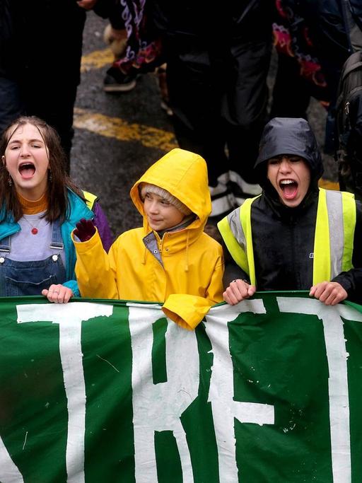 Greta Thunberg hält mit jungen Demonstrierenden ein grünes Banner. Die anderen Jugendlichen rufen etwa. Thunberg schaut zur Seite und winkt.