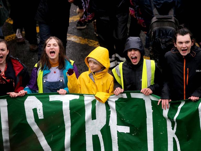 Greta Thunberg hält mit jungen Demonstrierenden ein grünes Banner. Die anderen Jugendlichen rufen etwa. Thunberg schaut zur Seite und winkt.