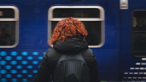 Eine rothaarige Frau steht mit dem Rücken zum Fotografen auf einem Bahnsteig, dahinter ein blauer Zug