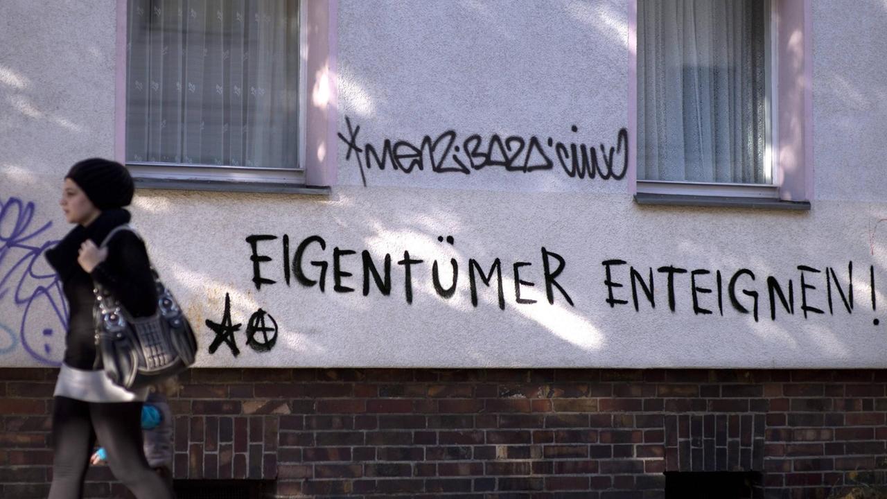 An einer Hauswand steht die Aufforderung Eigentümer zu enteignen. Eine Frau mit Kopftuch und Handtasche geht an dem Haus vorbei. An einer Hauswand steht die Aufforderung Eigentümer zu enteignen. Eine Frau mit Kopftuch und Handtasche geht an dem Haus vorbei.