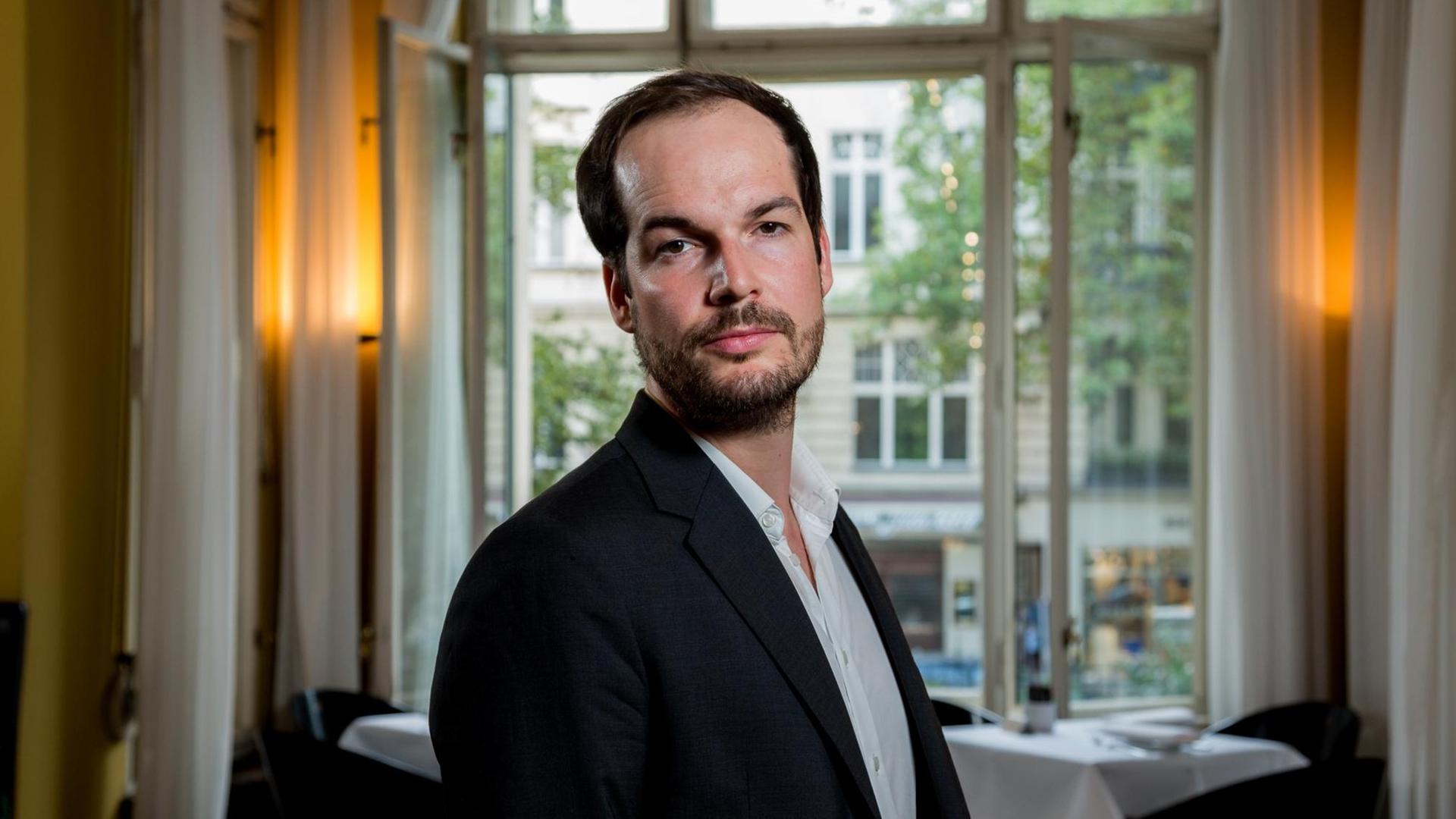 Max Czollek steht vor einem geöffneten Fenster und schaut in die Kamera Max Czollek steht vor einem geöffneten Fenster und schaut in die Kamera