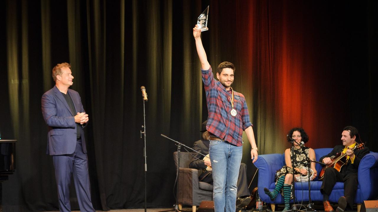 Der Kabarettist Nektarios Vlachopoulos steht mit einer Medaille auf der Bühne und hält eine Trophäe in die Höhe. Im Hintergrund links der Moderator des Abends Lars Reichow, rechts zwei Teilnehmer auf einer Couch.