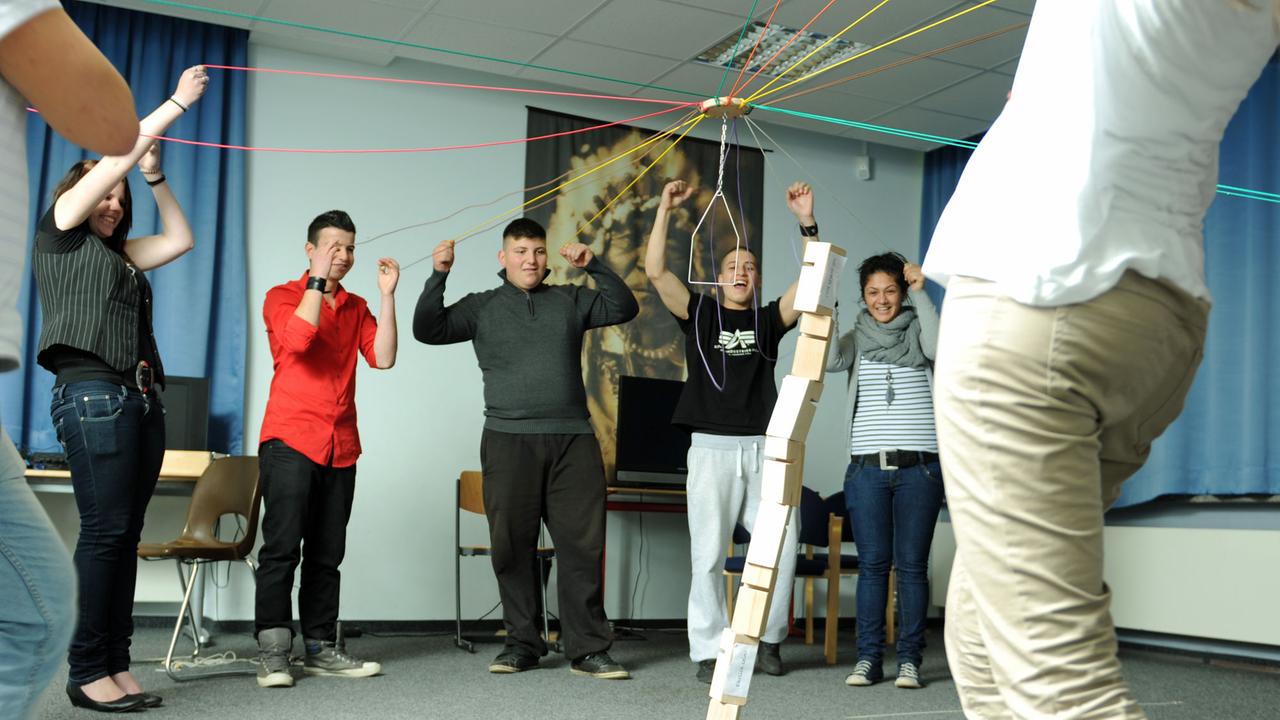 Eine Gruppe Schüler steht in einem Halbkreis vor einem Turm aus Holzklötzen, den sie mit einem von Schnüren gesteuerten Metallkran aufbauen. Eine Gruppe Schüler steht in einem Halbkreis vor einem Turm aus Holzklötzen, den sie mit einem von Schnüren gesteuerten Metallkran aufbauen.