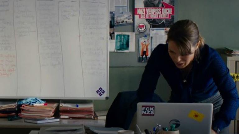 Ein Screenshot aus der Polizeiruf 110 - Folge "Für Janina": Anneke Kim Sarnau tippt auf ihrem Laptop, auf dem ein "FCK NZS"-Aufkleber und ein Aufkleber der Anti-Atombewegung kleben. Im Hintergrund unter anderem ein Poster mit der Aufschrift "Nazi verpiss Dich".