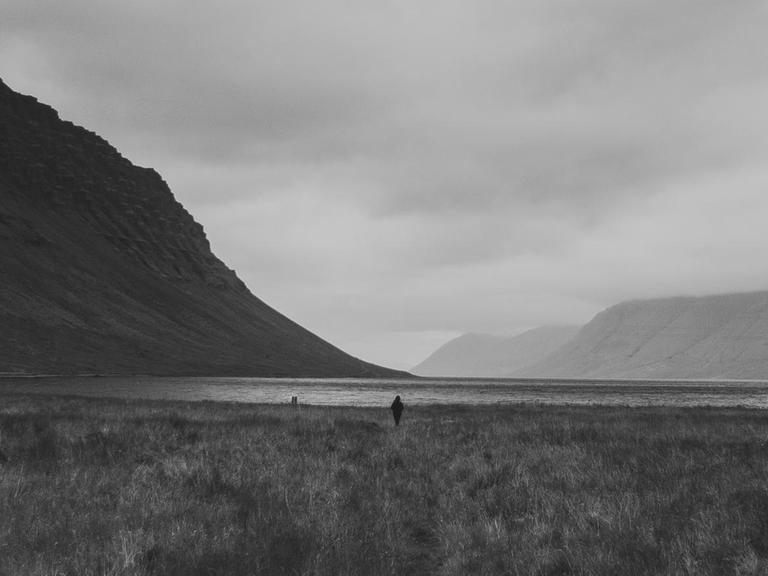 SW-Foto einer isländischen Landschaft mit Berg und See, nahe des Wasserfalls Dynjandi. In der Bildmitte ist die Silhouette eines Menschen zu sehen.