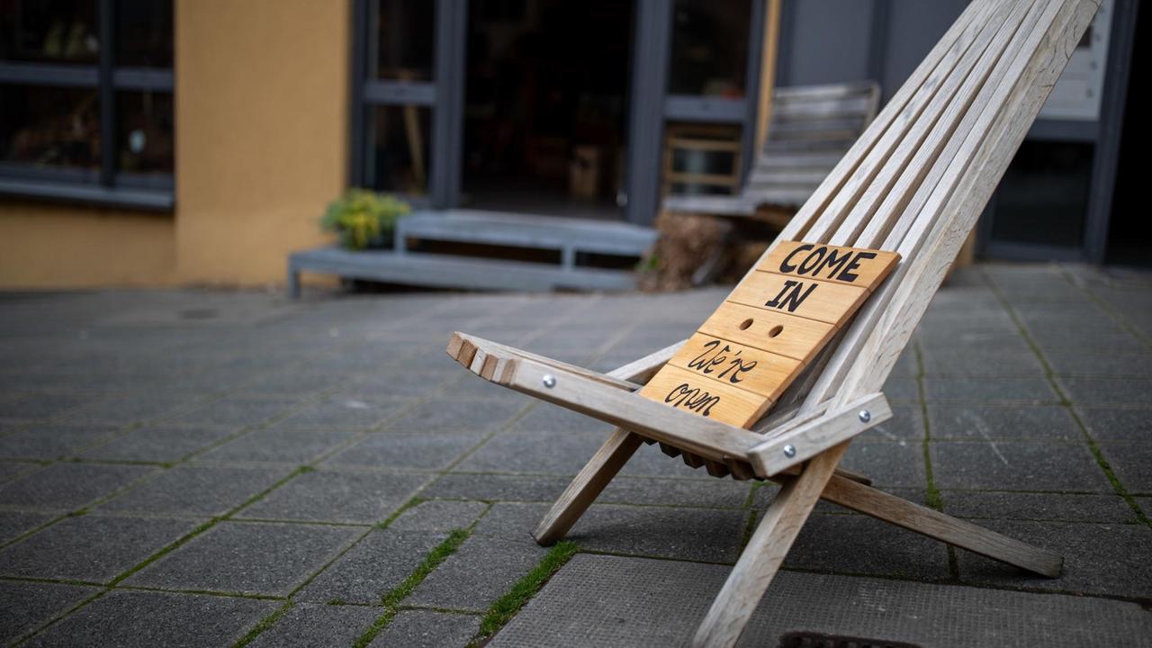"Come in - We're open" steht auf einem Schild aus Holz, welches vor einem Geschäft in der Innenstadt auf einem Liegestuhl steht. "Come in - We're open" steht auf einem Schild aus Holz, welches vor einem Geschäft in der Innenstadt auf einem Liegestuhl steht.