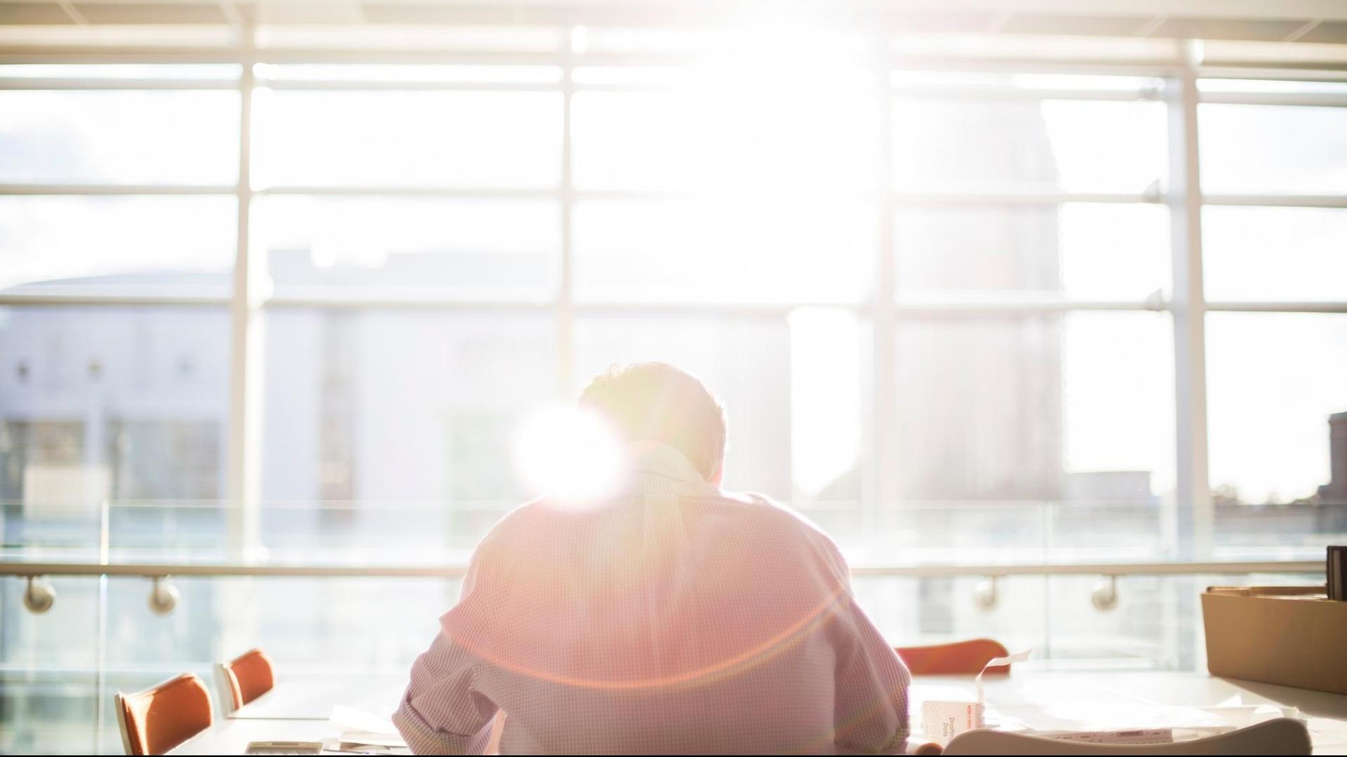 Ein Mann sitzt an einem Konferenztisch mit dem Gesicht zur Fensterfront, durch die die Sonne scheint. Ein Mann sitzt an einem Konferenztisch mit dem Gesicht zur Fensterfront, durch die die Sonne scheint.