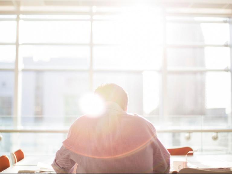Ein Mann sitzt an einem Konferenztisch mit dem Gesicht zur Fensterfront, durch die die Sonne scheint.