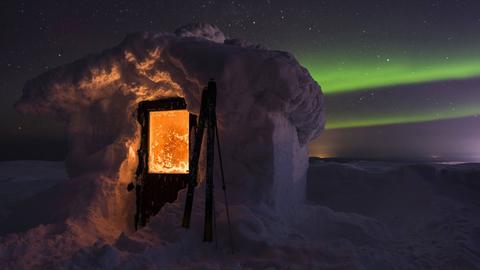 Eine mit Schnee bedeckte Hütte, aus der von innen Licht scheint. Im Hintergrund Polarlichter.