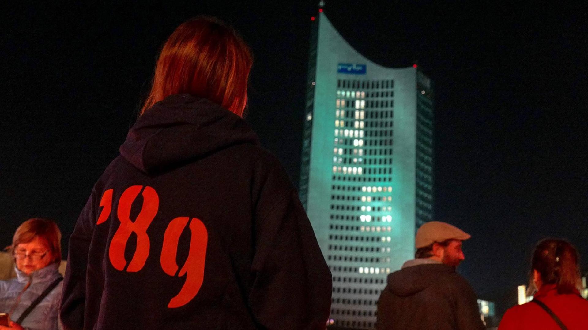 Eine junge Frau steht mit einem Pulli, auf dem die Aufschrift "89" zu lesen ist, vor dem City-Hochhaus in Leipzig.