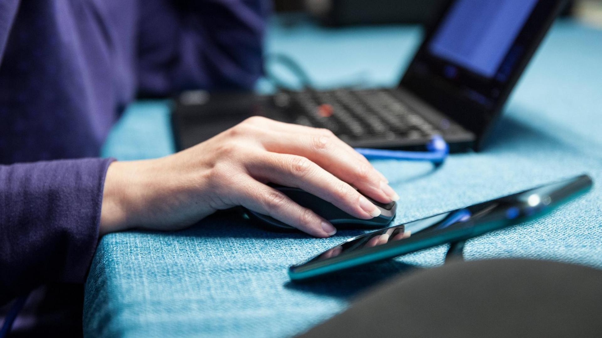 Eine Person sitzt an einem Tisch im Homeoffice an einem Laptop, die Hand liegt auf der Maus, daneben steht ein Handy. Eine Person sitzt an einem Tisch im Homeoffice an einem Laptop, die Hand liegt auf der Maus, daneben steht ein Handy.