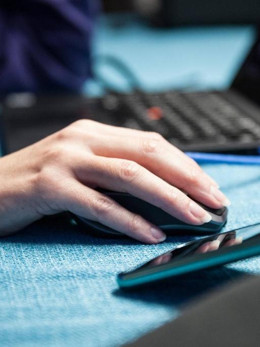 Eine Person sitzt an einem Tisch im Homeoffice an einem Laptop, die Hand liegt auf der Maus, daneben steht ein Handy.