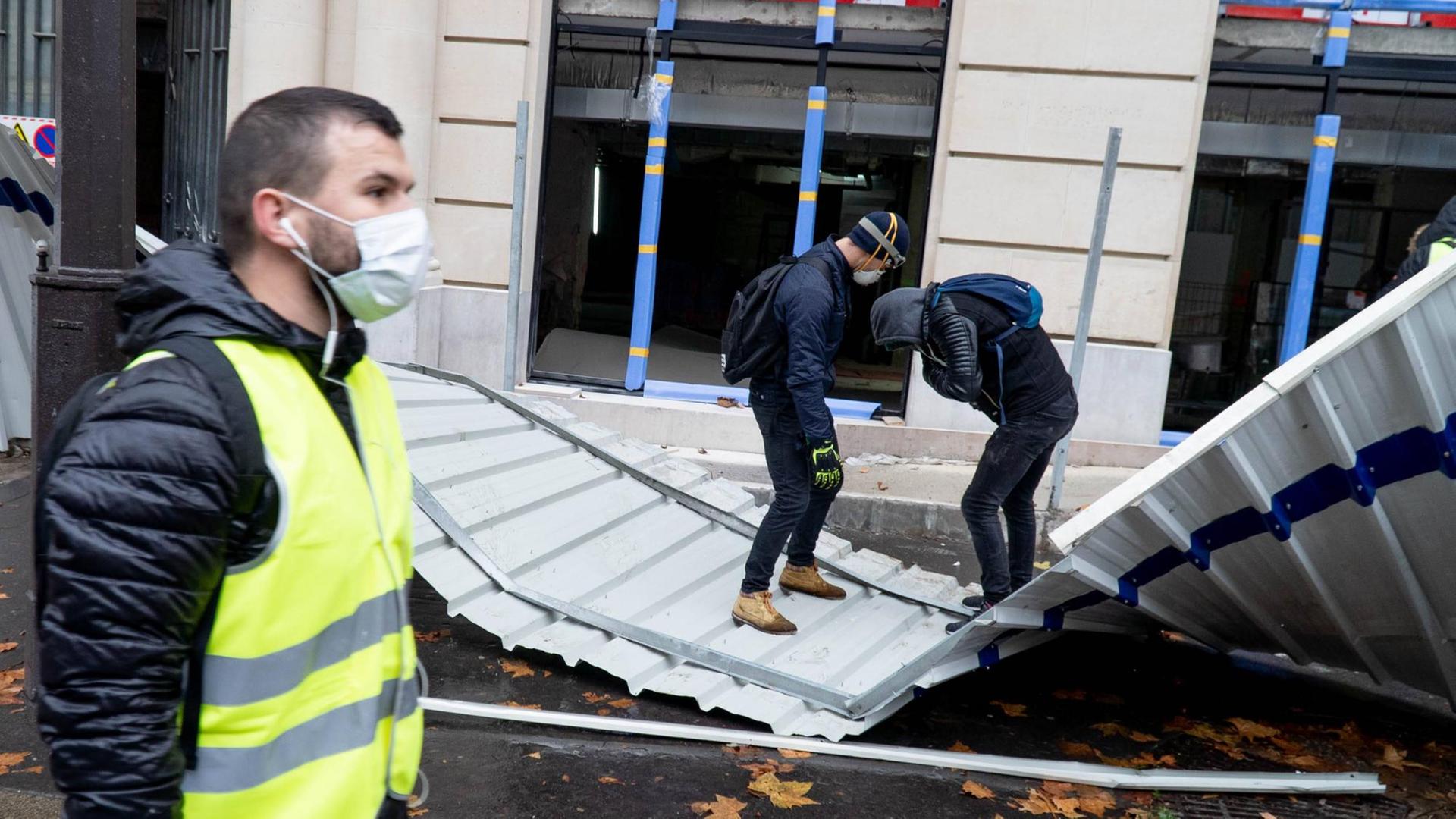 Ein Mann in einer gelben Weste steht im Bildvordergrund, während zwei Männer im Hintergrund eine Absperrung zerstören Ein Mann in einer gelben Weste steht im Bildvordergrund, während zwei Männer im Hintergrund eine Absperrung zerstören