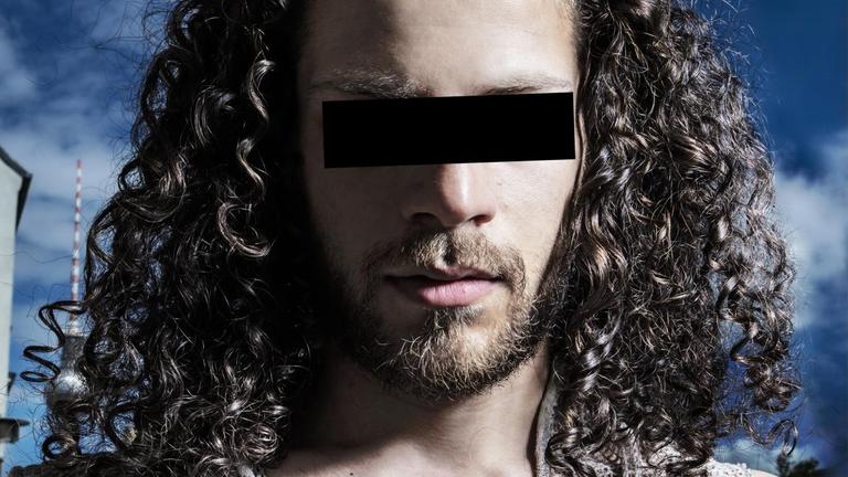 Ein Mann mit langen Haaren hat einen schwarzen Balken vor den Augen, um unkenntlich zu bleiben.