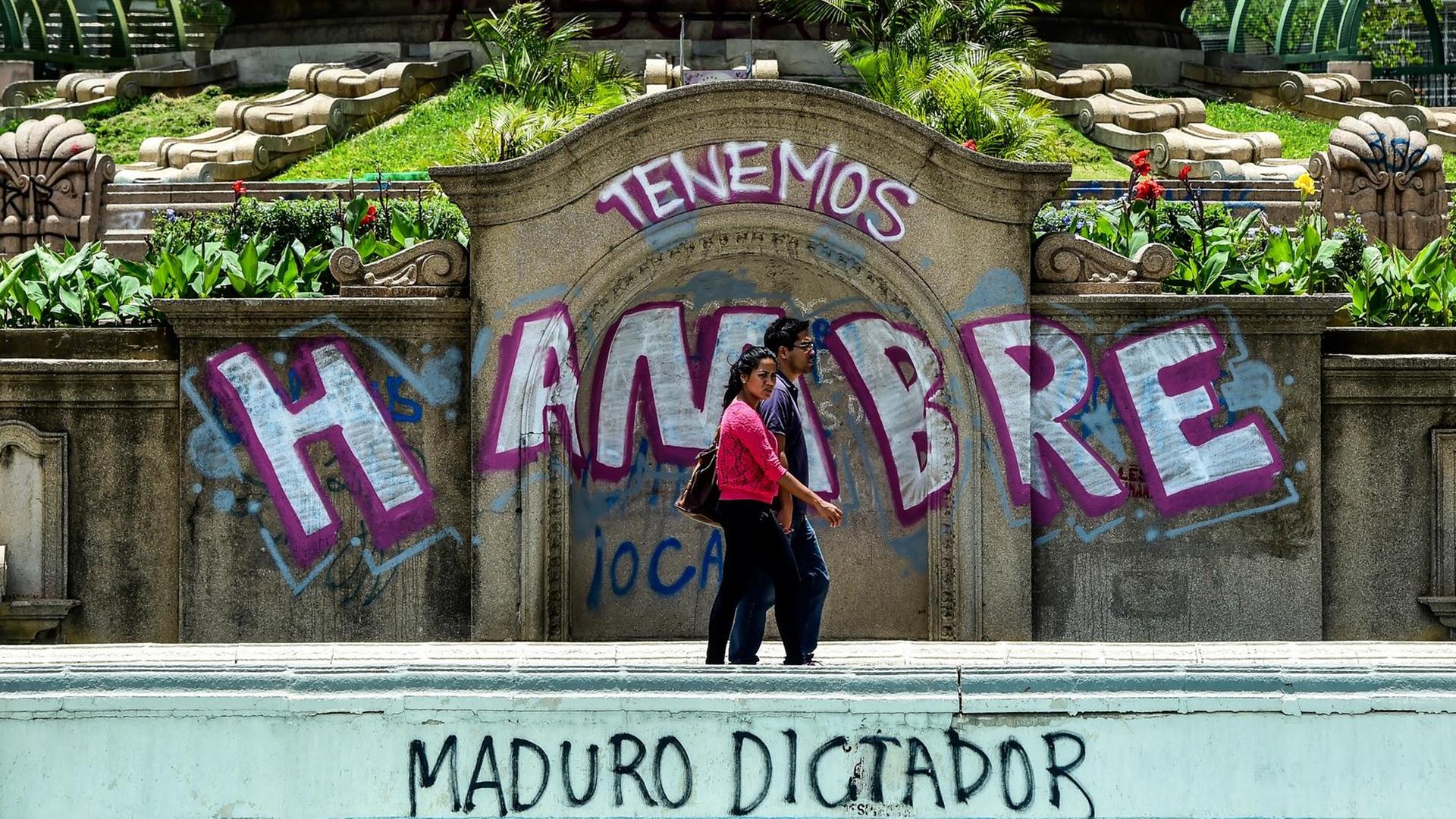 Ein Paar läuft auf der Straße. Dahinter zu sehen: Graffiti mit dem Text "Wir haben Hunger" und "Diktator Maduro" in Venezuelas Hauptstadt Caracas. Ein Paar läuft auf der Straße. Dahinter zu sehen: Graffiti mit dem Text "Wir haben Hunger" und "Diktator Maduro" in Venezuelas Hauptstadt Caracas.