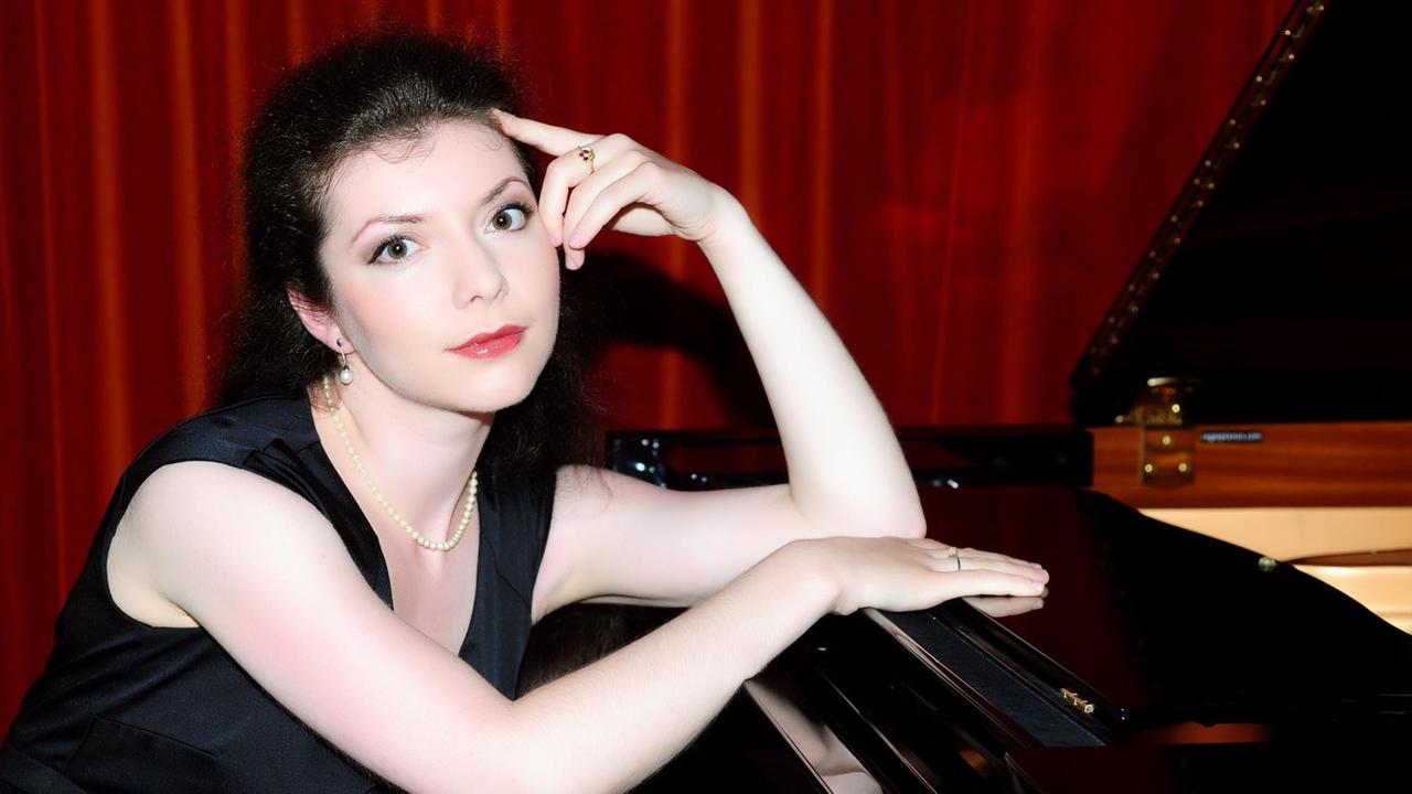 Die Pianistin sitzt an einem Flügel vor rotem Vorhang. Dabei hat sie einen Arm auf das Notenpult gestütz. Die Pianistin sitzt an einem Flügel vor rotem Vorhang. Dabei hat sie einen Arm auf das Notenpult gestütz.