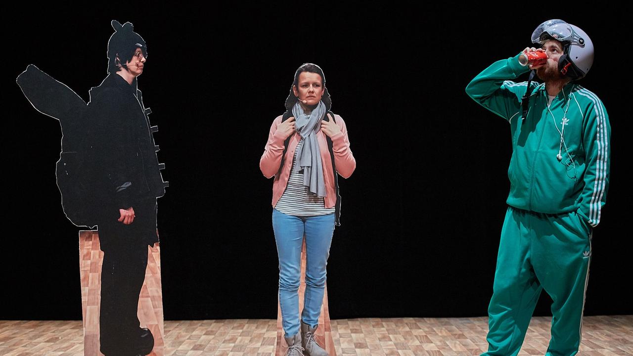 Ein Schauspieler steht auf der Bühne, er trägt einen grünen Trainingsanzug. Dazu sind noch zwei Pappfiguren aufgestellt. Ein Schauspieler steht auf der Bühne, er trägt einen grünen Trainingsanzug. Dazu sind noch zwei Pappfiguren aufgestellt.