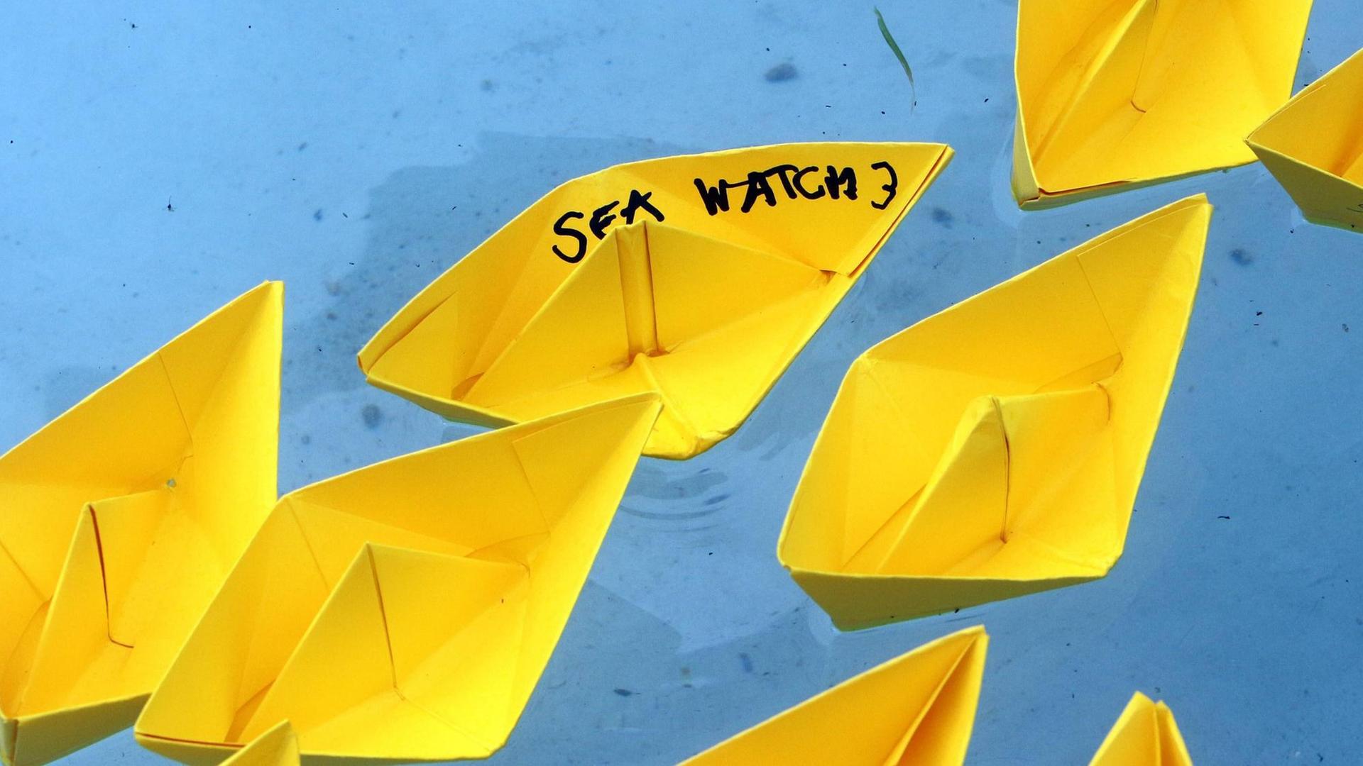 Gelbe Papierschiffchen, die Flüchtlingsboote auf dem Mittelmeer darstellen sollen, wurden von Demonstranten in das Wasser des Brunnens am Schlossplatz gesetzt. Ein Schiffchen trägt die Aufschrift Sea Watch 3. Gelbe Papierschiffchen, die Flüchtlingsboote auf dem Mittelmeer darstellen sollen, wurden von Demonstranten in das Wasser des Brunnens am Schlossplatz gesetzt. Ein Schiffchen trägt die Aufschrift Sea Watch 3.
