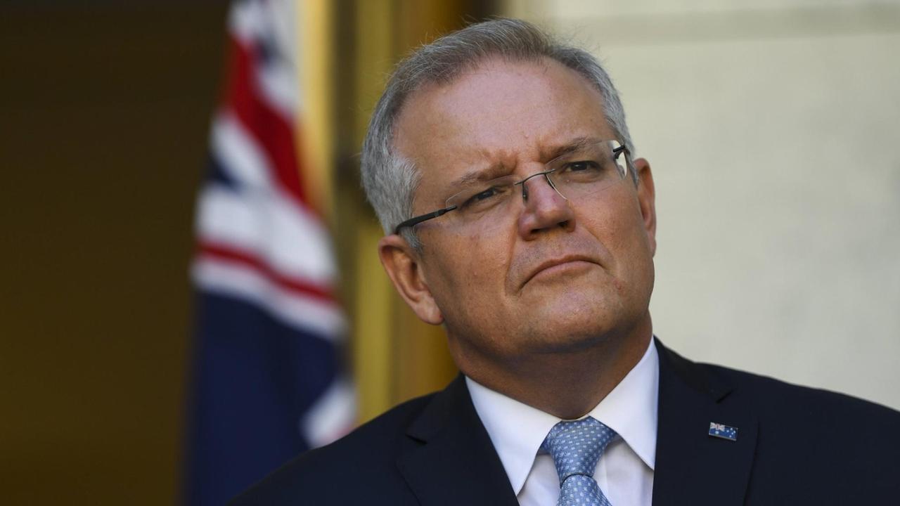 Ein Mann mit Brille, Krawatte und Anzug steht vor der australischen Flagge und schaut kritisch.