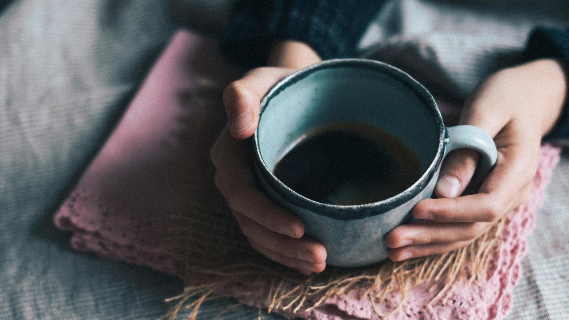 Zwei Hände, die auf einer Decke liegen umfassen eine Tasse mit dunkler Flüssigkeit Zwei Hände, die auf einer Decke liegen umfassen eine Tasse mit dunkler Flüssigkeit