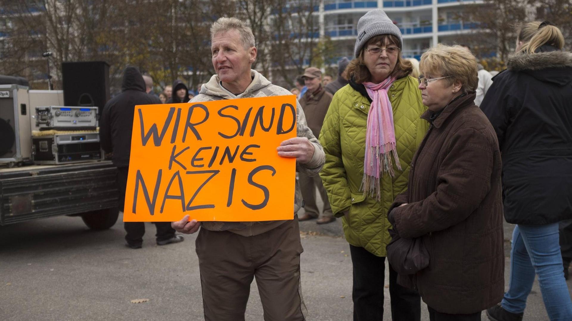 Auf dem Foto zu sehen sind drei ältere Personen auf einer Demonstration gegen die geplante Unterbringung von Asylbewerbern in Marzahn-Hellersdorf im Herbst 2014. Ein Mann trägt ein Schild mit der Aufschrift: "Wir sind keine Nazis". 
