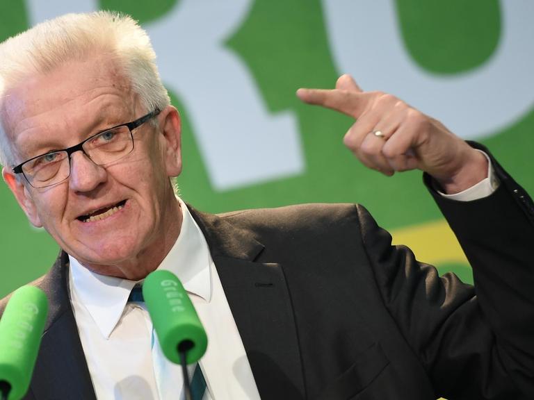 Sie sehen Baden-Württembergs Ministerpräsident Kretschmann, er spricht in zwei grüne Mikrofone.