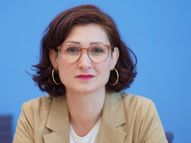 Ferda Ataman von den Neue deutsche Medienmacher*innen schaut mit Brille vor blauem Hintergrund in die Kamera.