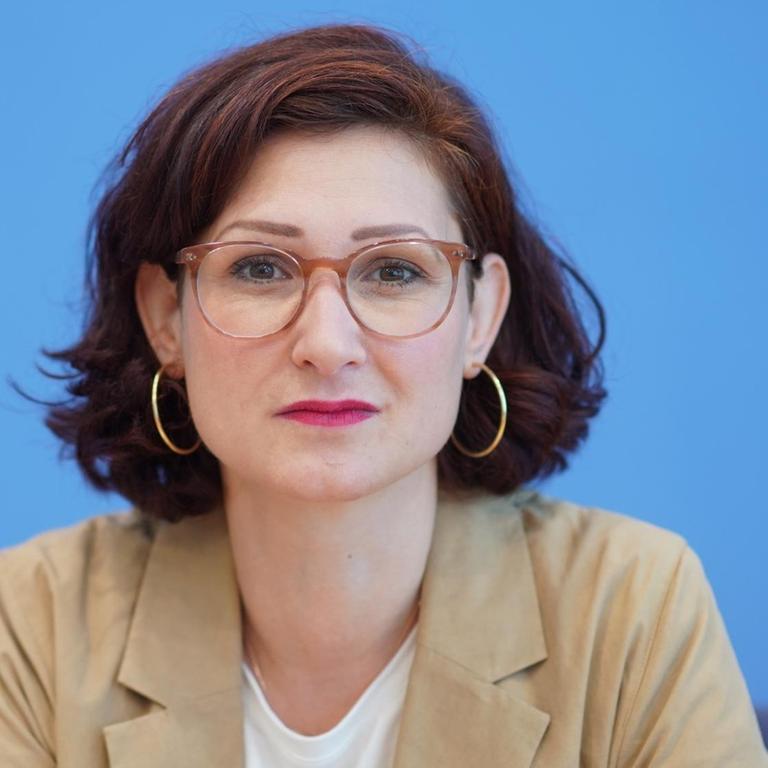Ferda Ataman von den Neue deutsche Medienmacher*innen schaut mit Brille vor blauem Hintergrund in die Kamera.