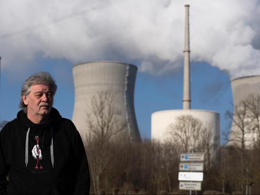 Ein Mann mit grauen Haaren und Schnauzbart steht im schwarzen Kapuzenshirt vor einem Atomkraftwerk, aus dem weißer Rauch in den blauen Himmel aufsteigt.